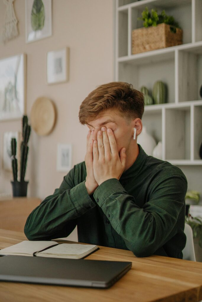 A man in green shirt looking frustrated, covering his face with hands at a desk.